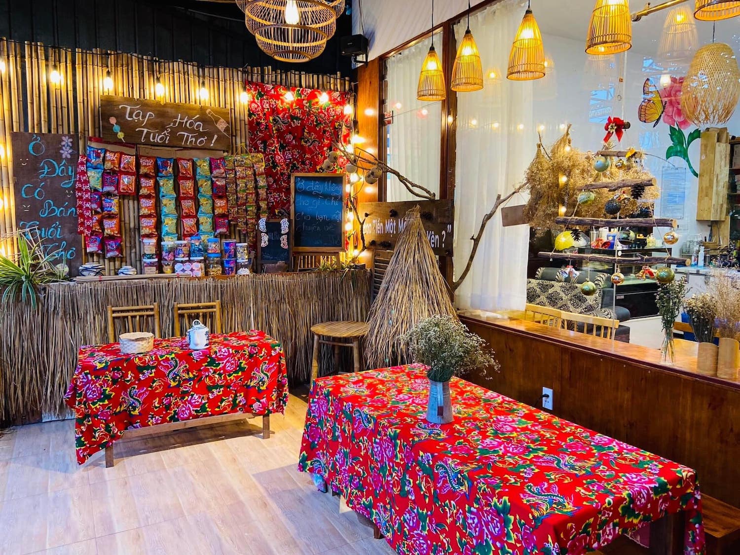 Không gian quán nhỏ với bàn ghế tre, vải trải bàn hoa văn, trưng bày nhiều gói bánh kẹo và trang trí đèn lồng.