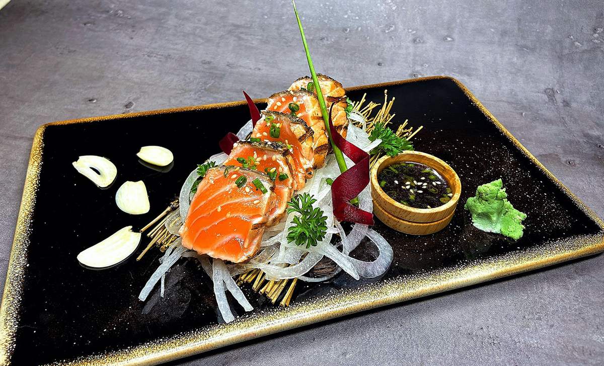 Đĩa sushi c&aacute; hồi trang tr&iacute; đẹp mắt với h&agrave;nh l&aacute;, tỏi, v&agrave; nước chấm, b&agrave;y tr&ecirc;n đĩa đen sang trọng.