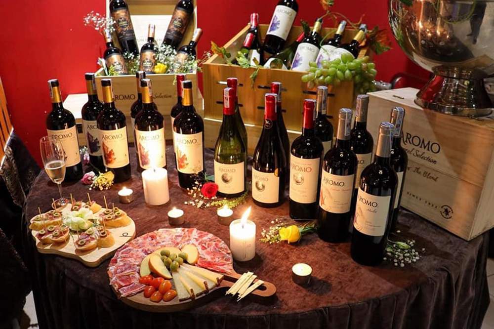 B&agrave;n tiệc với nhiều chai rượu vang, nến, đồ ăn nhẹ, trang tr&iacute; hoa v&agrave; tr&aacute;i c&acirc;y tr&ecirc;n khăn trải b&agrave;n m&agrave;u n&acirc;u.