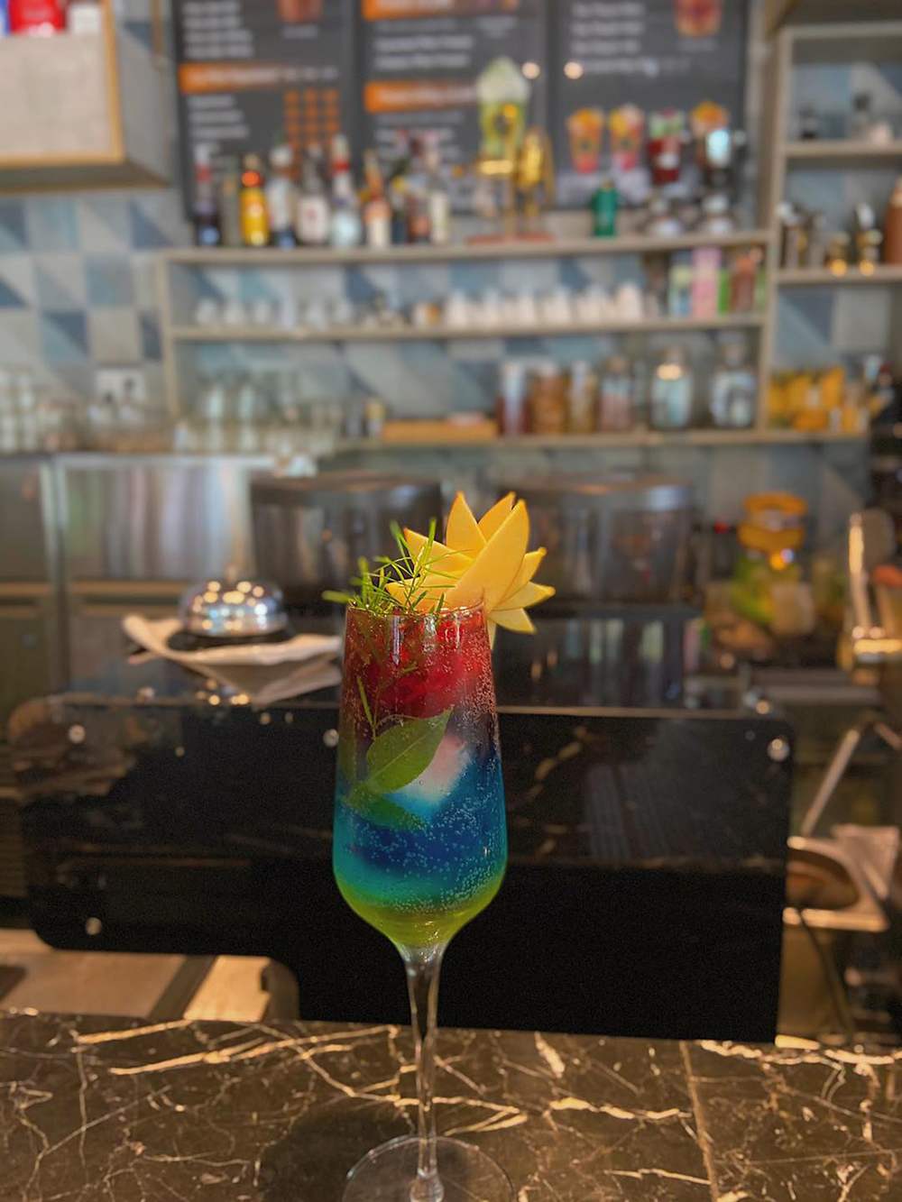 Ly cocktail nhiều m&agrave;u sắc với trang tr&iacute; tr&aacute;i c&acirc;y tr&ecirc;n b&agrave;n quầy bar, nền l&agrave; kệ đựng chai lọ v&agrave; cốc ch&eacute;n.