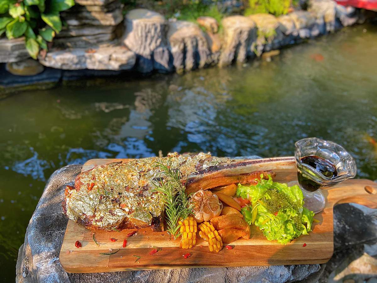 M&oacute;n thịt nướng phủ v&agrave;ng tr&ecirc;n thớt gỗ, trang tr&iacute; với rau củ, b&ecirc;n cạnh l&agrave; ch&eacute;n sốt, hậu cảnh l&agrave; hồ nước v&agrave; c&acirc;y xanh.