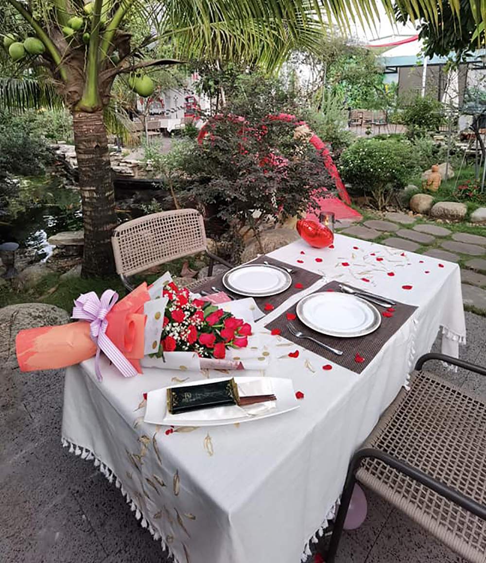 B&agrave;n ăn ngo&agrave;i trời trang tr&iacute; hoa hồng đỏ, khăn trải b&agrave;n trắng, dưới b&oacute;ng c&acirc;y dừa, cạnh khu vườn xanh m&aacute;t.