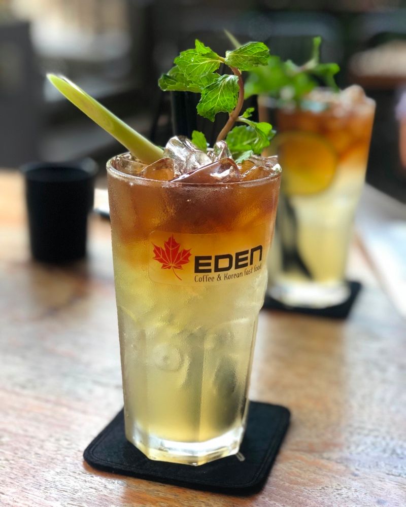 Ly trà đá trong suốt trên bàn gỗ, trang trí bằng lá bạc hà và sả, có logo quán Eden.