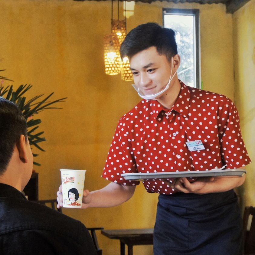 Nhân viên phục vụ mặc áo chấm bi đỏ, đeo khẩu trang, đưa cốc giấy cho khách trong quán cà phê ấm cúng.
