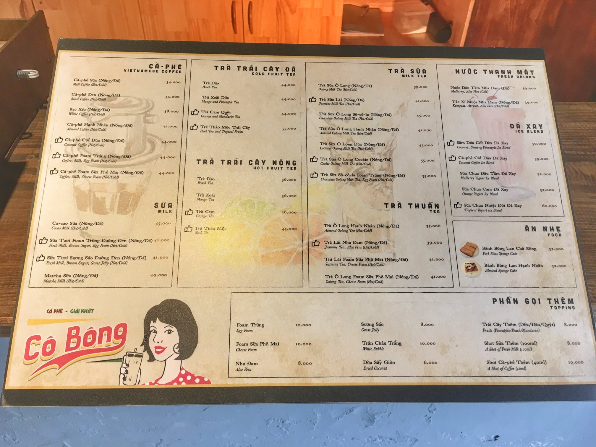 Menu đồ uống và món ăn trên bàn gỗ, bao gồm cà phê, trà sữa, nước thanh mát và đồ ăn nhẹ.