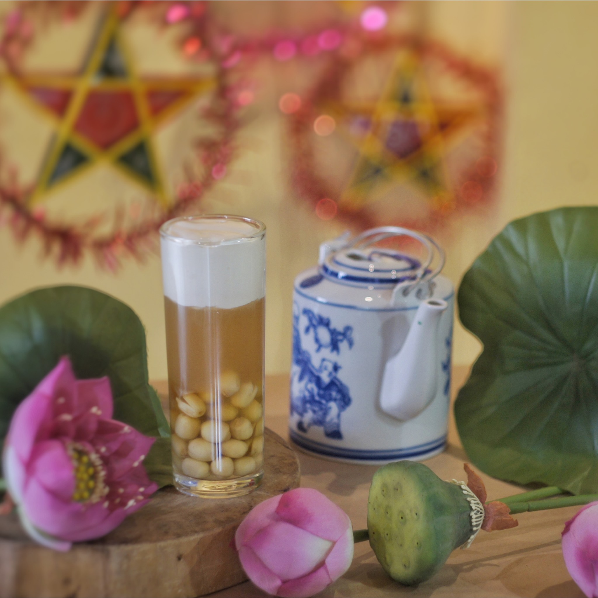 Ly sữa đậu và ấm trà trên bàn, xung quanh có hoa sen và lá sen, nền trang trí đèn ông sao.
