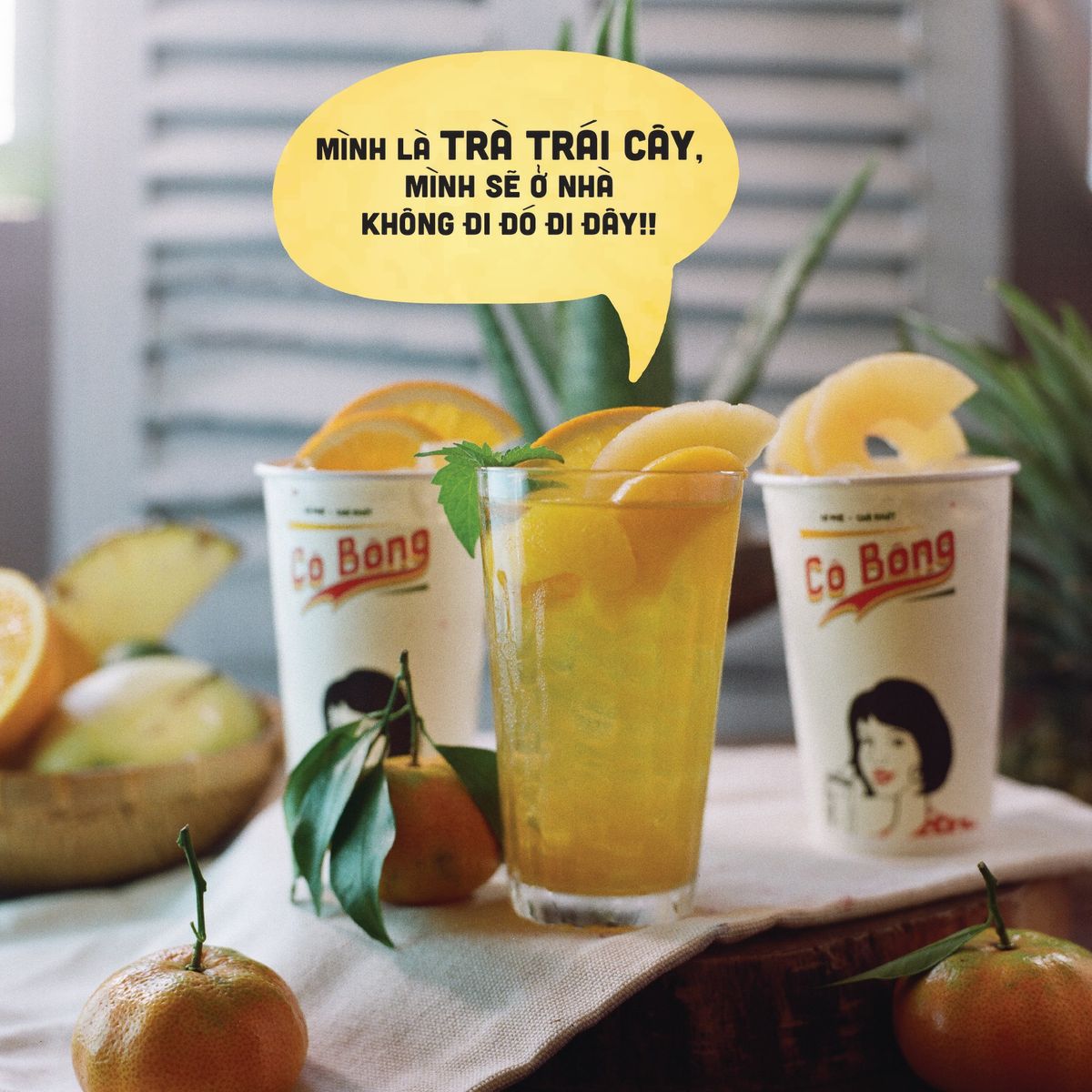 Ly trà trái cây và hai ly nhựa trên bàn, xung quanh là trái cây. Bảng chữ thoại màu vàng nổi bật.
