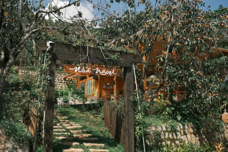 Cổng gỗ dẫn lối vào nhà gỗ nhỏ trong vườn xanh mát, khung cảnh yên bình và gần gũi với thiên nhiên.