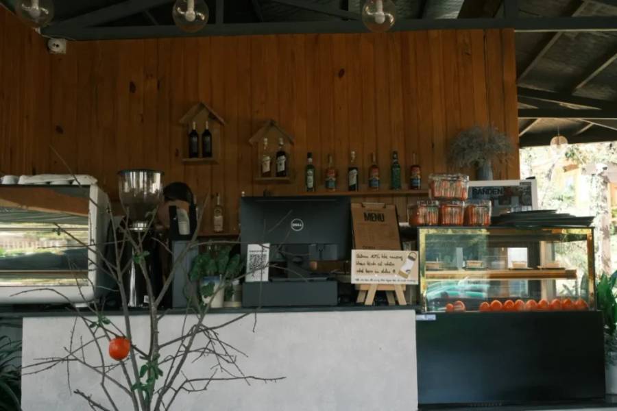 Quầy bar với m&aacute;y pha c&agrave; ph&ecirc;, đồ uống tr&ecirc;n kệ v&agrave; tủ k&iacute;nh trưng b&agrave;y cam ở ph&iacute;a trước.