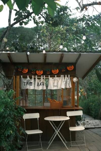 Quầy b&aacute;n b&aacute;nh nhỏ trong vườn, trang tr&iacute; đ&egrave;n v&agrave; b&iacute; ng&ocirc; Halloween, c&oacute; b&agrave;n ghế trắng ph&iacute;a trước.