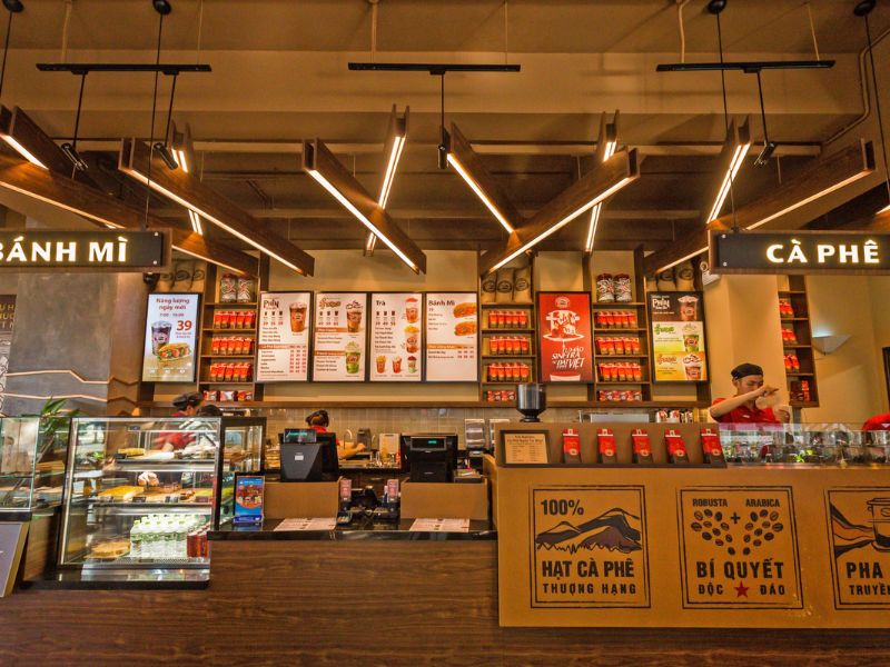 Quầy phục vụ trong qu&aacute;n c&agrave; ph&ecirc;, với bảng menu, m&aacute;y t&iacute;nh tiền v&agrave; c&aacute;c sản phẩm b&agrave;y tr&ecirc;n kệ.