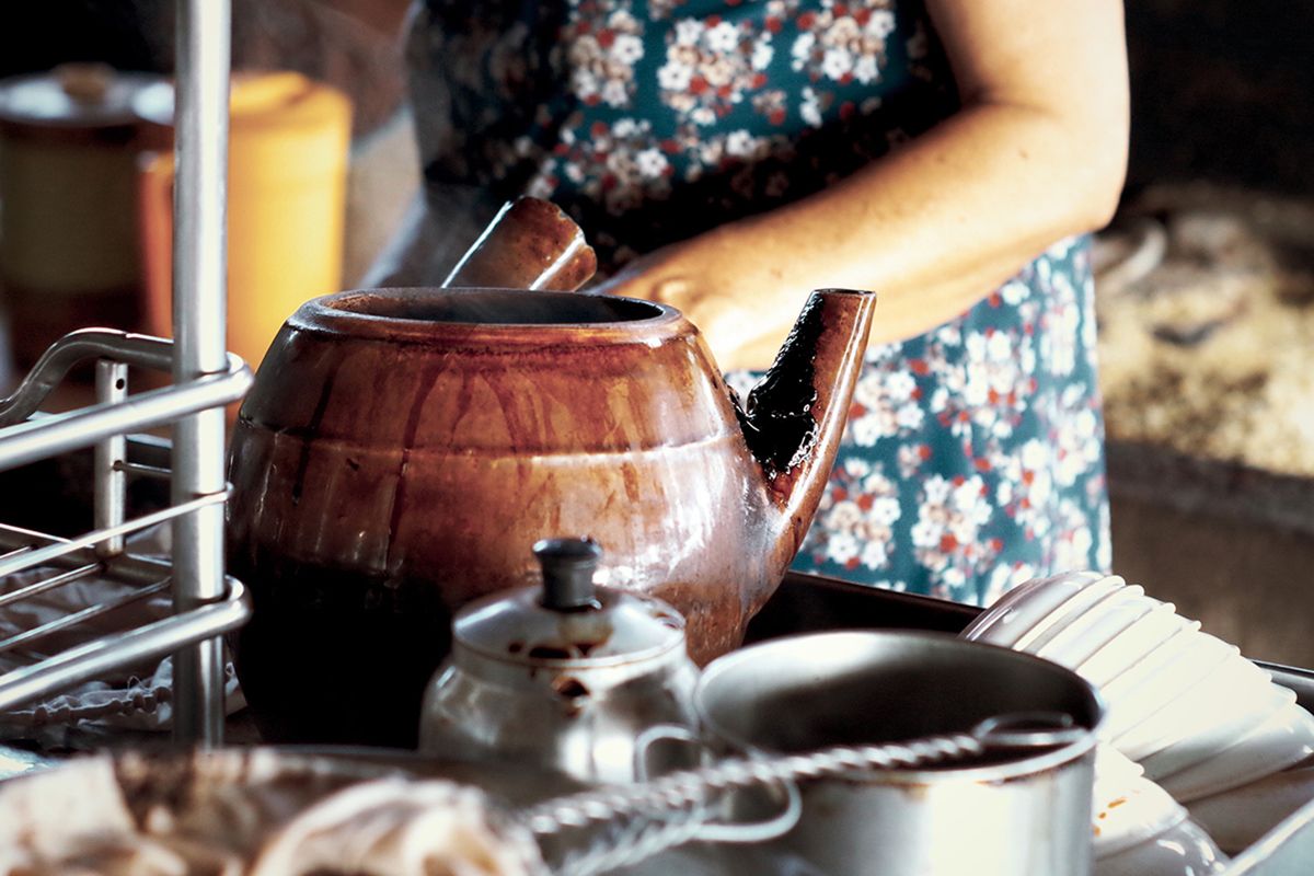 Ấm đất v&agrave; dụng cụ nh&agrave; bếp tr&ecirc;n b&agrave;n, với người mặc v&aacute;y hoa đứng ph&iacute;a sau.