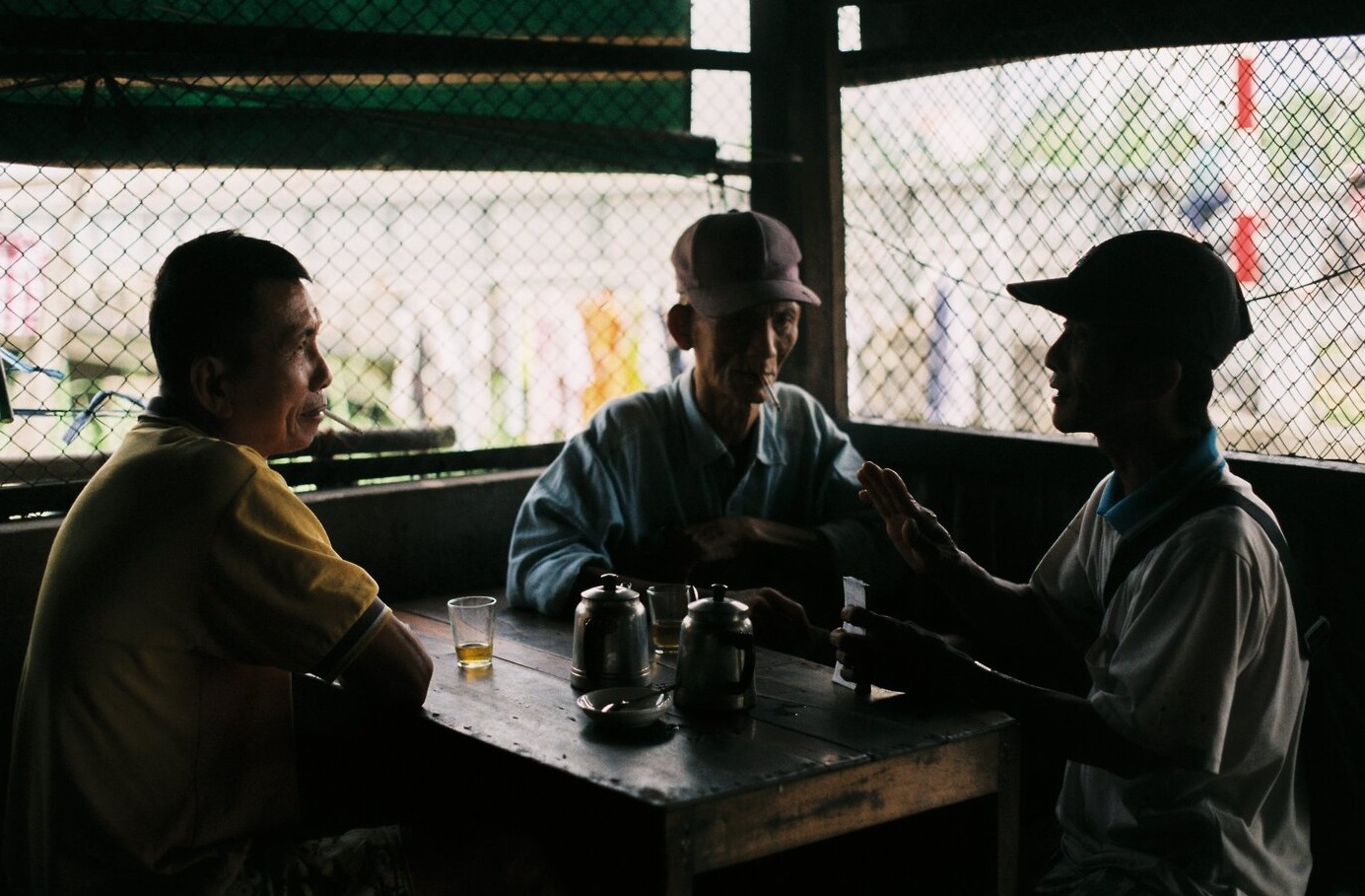 Ba người ngồi quanh b&agrave;n trong qu&aacute;n c&agrave; ph&ecirc; tối, tr&ograve; chuyện c&ugrave;ng nhau, với ấm tr&agrave; v&agrave; ly nhỏ tr&ecirc;n b&agrave;n.