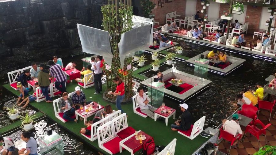 Qu&aacute;n c&agrave; ph&ecirc; với b&agrave;n ghế đặt tr&ecirc;n mặt nước, kh&aacute;ch ngồi thưởng thức đồ uống trong kh&ocirc;ng gian tho&aacute;ng đ&atilde;ng, c&oacute; c&acirc;y xanh.