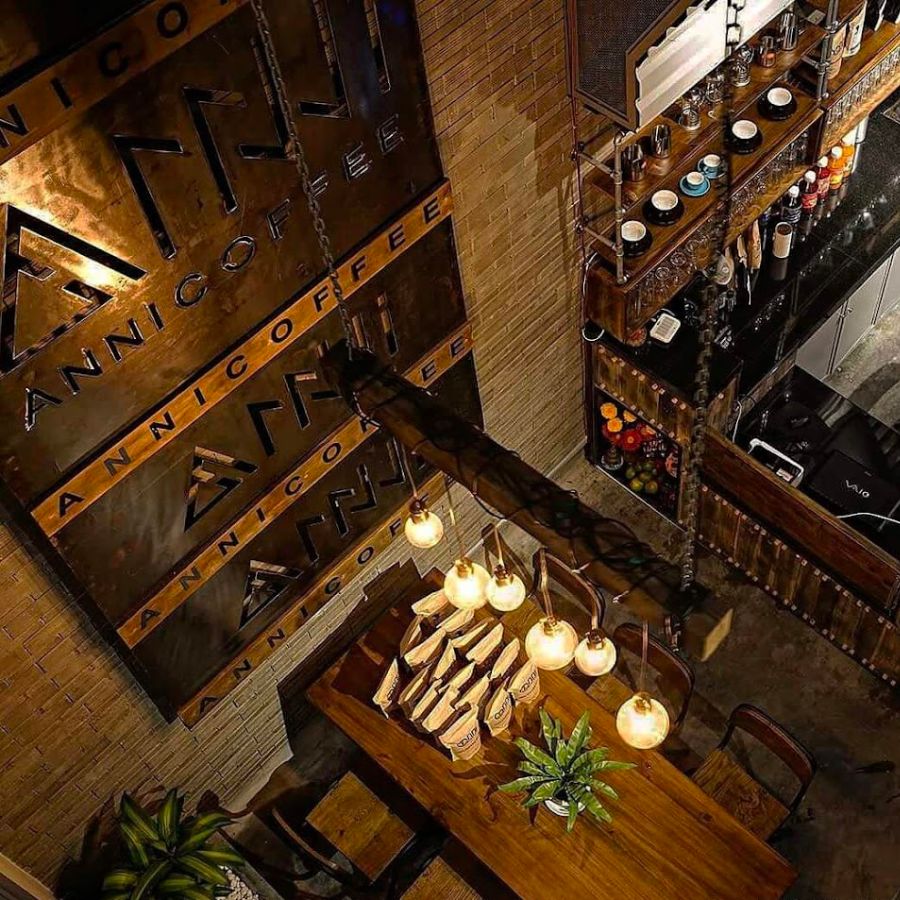 Qu&aacute;n c&agrave; ph&ecirc; với b&agrave;n gỗ, đ&egrave;n treo, kệ chứa đồ uống v&agrave; tường c&oacute; logo lớn, kh&ocirc;ng gian ấm c&uacute;ng.