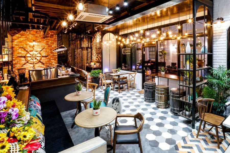 Qu&aacute;n c&agrave; ph&ecirc; với nội thất ấm c&uacute;ng, b&agrave;n ghế gỗ, trang tr&iacute; c&acirc;y xanh v&agrave; đ&egrave;n treo, tường gạch v&agrave; s&agrave;n l&aacute;t gạch hoa.