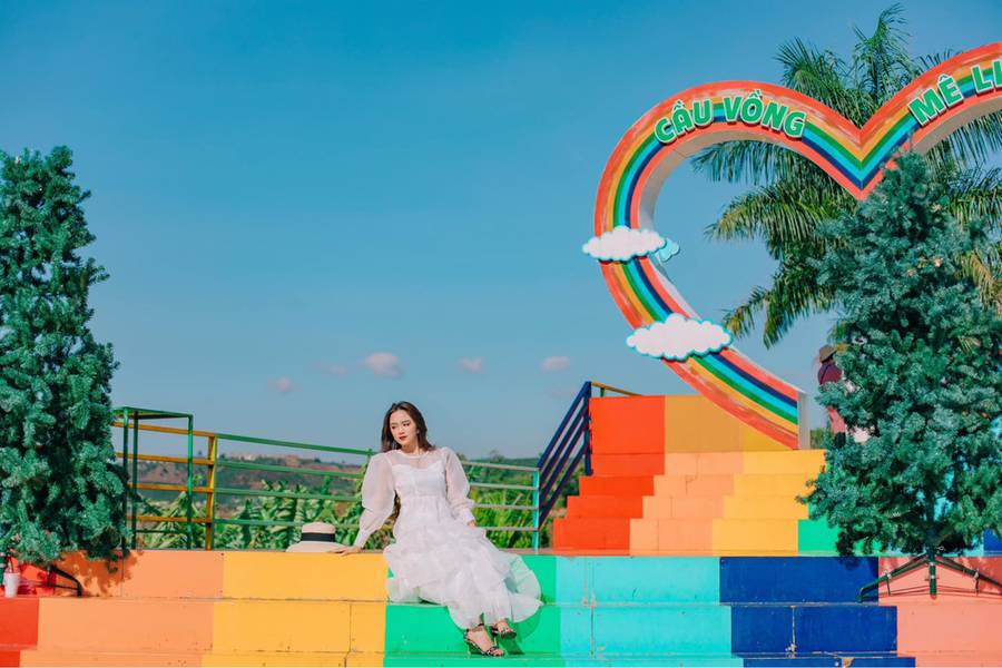 Người phụ nữ mặc v&aacute;y trắng ngồi tr&ecirc;n bậc thang nhiều m&agrave;u, xung quanh c&oacute; c&acirc;y v&agrave; cổng h&igrave;nh tr&aacute;i tim.