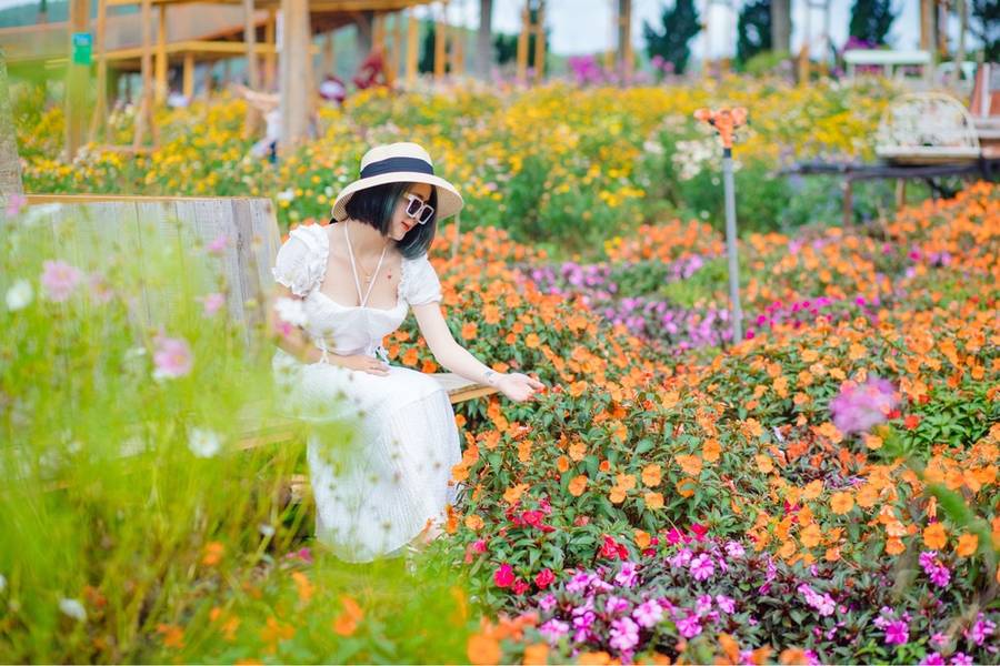 Người phụ nữ mặc v&aacute;y trắng ngồi tr&ecirc;n ghế trong vườn hoa đầy m&agrave;u sắc, đội mũ rộng v&agrave;nh, đeo k&iacute;nh r&acirc;m.