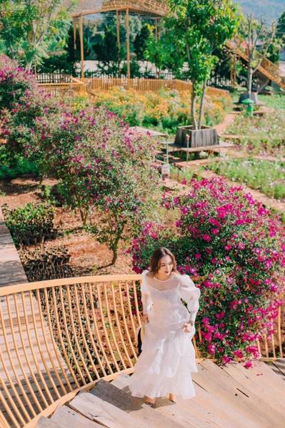 Người phụ nữ mặc v&aacute;y trắng đi tr&ecirc;n cầu gỗ, xung quanh l&agrave; c&acirc;y cối v&agrave; hoa m&agrave;u t&iacute;m trong khu vườn.