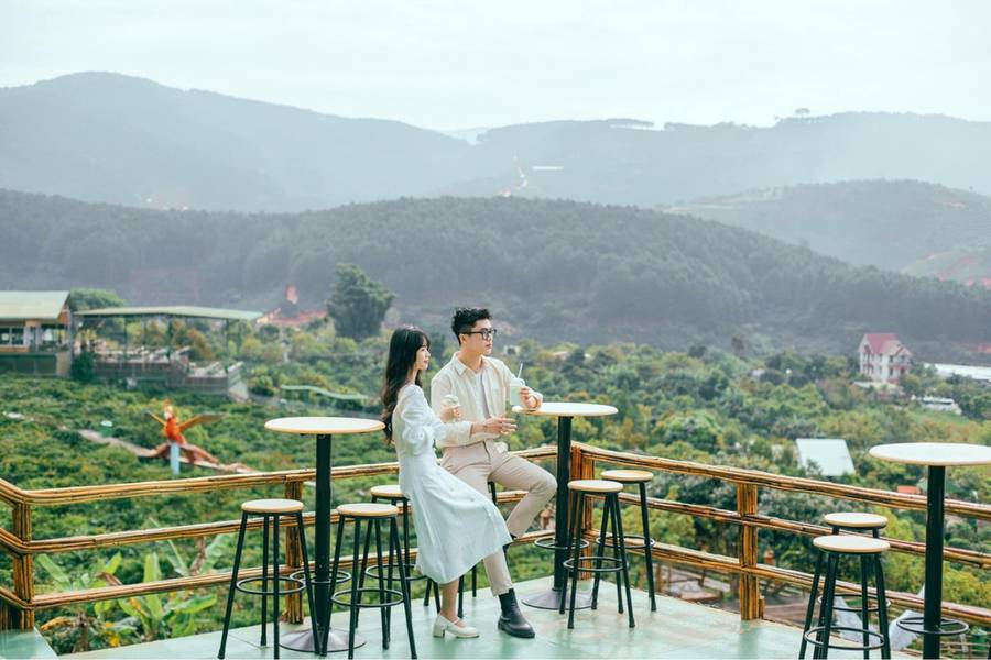 Hai người ngồi tr&ecirc;n ban c&ocirc;ng, thưởng thức đồ uống, ph&ocirc;ng nền l&agrave; khung cảnh n&uacute;i rừng xanh m&aacute;t.