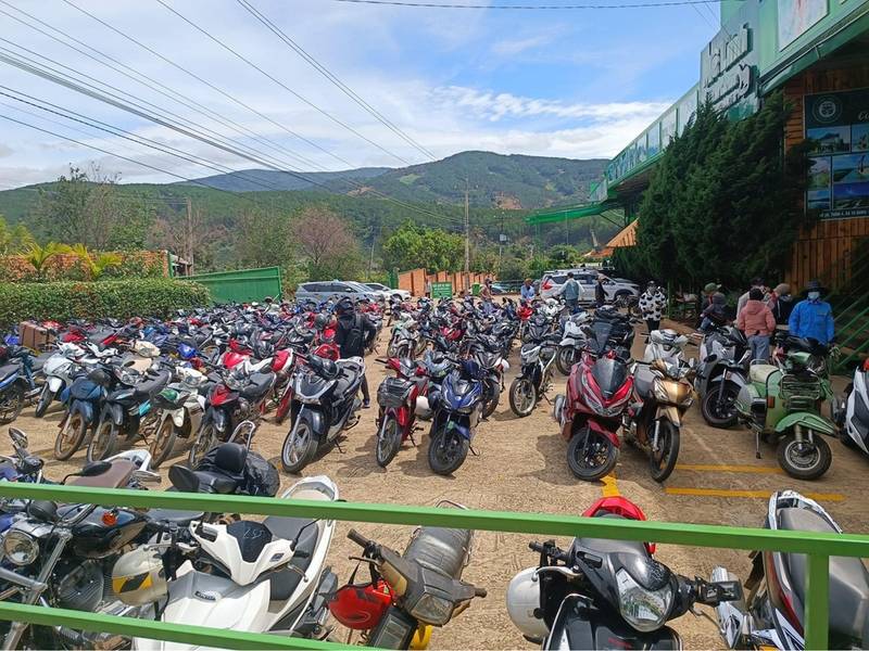 B&atilde;i đỗ xe m&aacute;y đ&ocirc;ng đ&uacute;c với nhiều xe m&aacute;y đa dạng m&agrave;u sắc, nền l&agrave; cảnh quan n&uacute;i non v&agrave; c&acirc;y xanh.