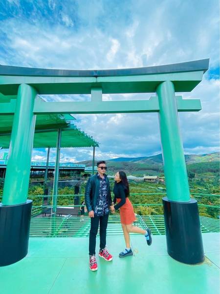 Hai người đứng dưới cổng truyền thống m&agrave;u xanh, bầu trời xanh v&agrave; n&uacute;i non ph&iacute;a sau.