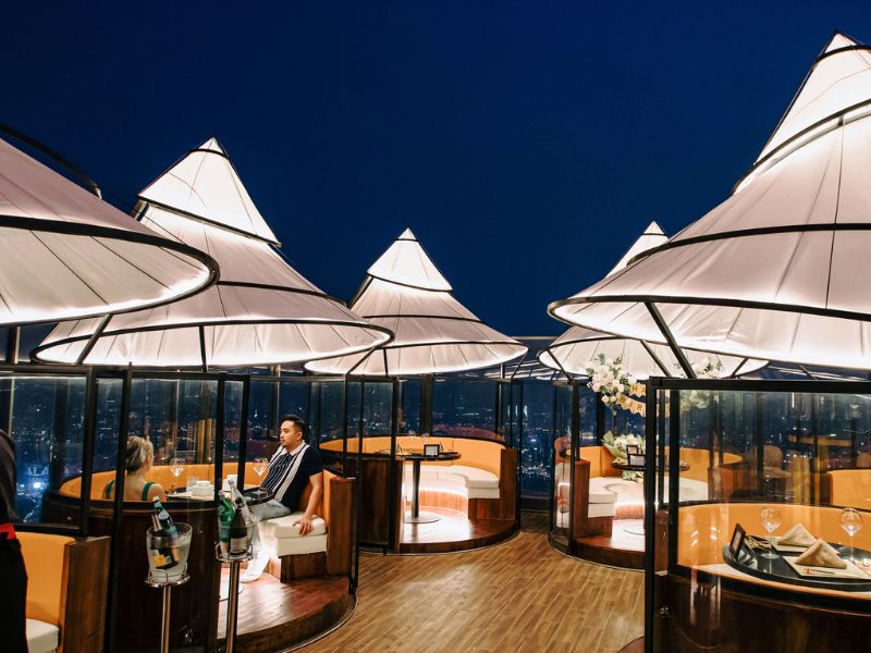 Nh&agrave; h&agrave;ng tr&ecirc;n cao với b&agrave;n ghế sang trọng, &aacute;nh đ&egrave;n ấm &aacute;p, trời tối, hai người ngồi tr&ograve; chuyện trong kh&ocirc;ng gian mở.