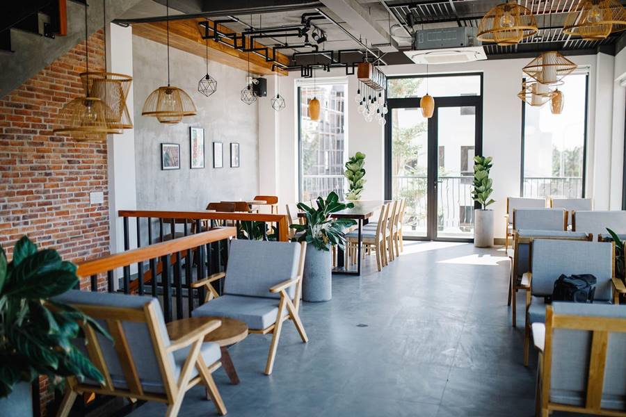 Quán cà phê nội thất hiện đại với bàn ghế gỗ, cây xanh, đèn treo và tường gạch, ánh sáng tự nhiên từ cửa sổ lớn.