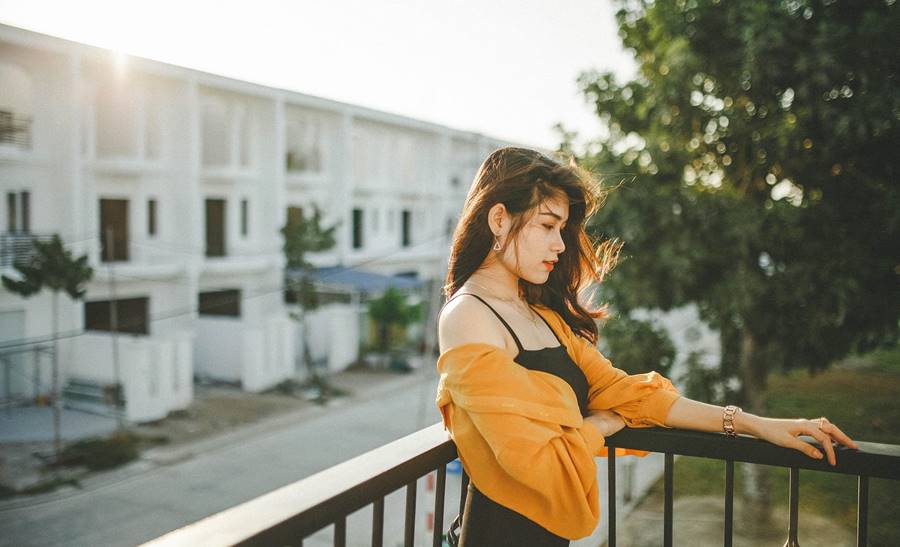 Người phụ nữ đứng tựa lan can, phía sau là dãy nhà trắng và cây xanh, ánh nắng chiếu sáng từ phía sau.
