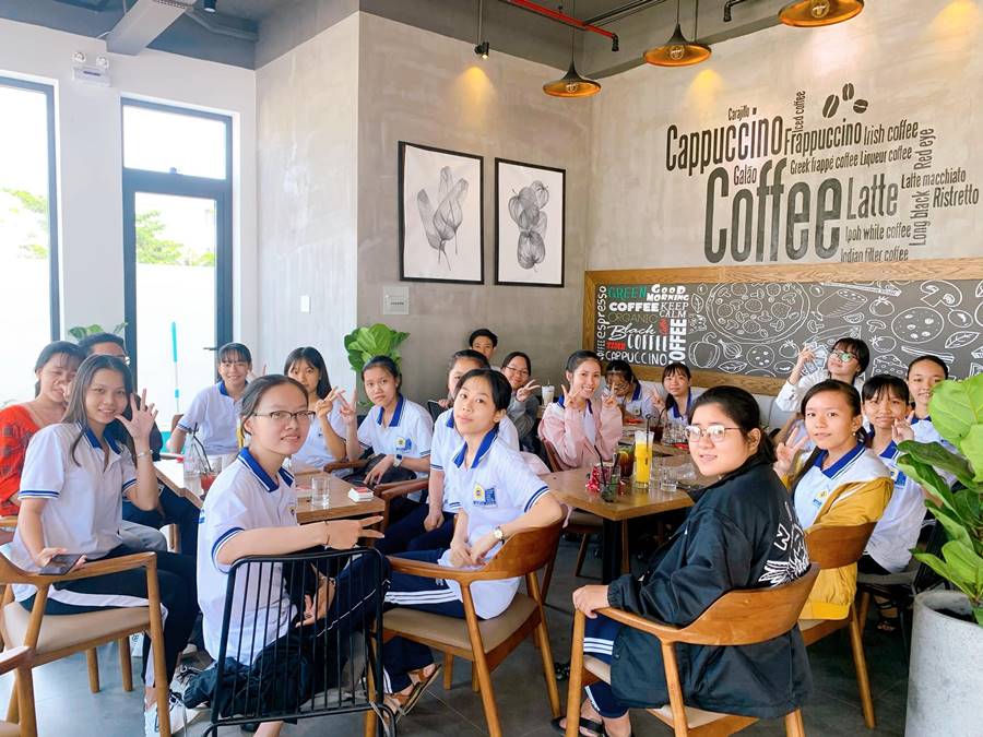 Một nhóm người ngồi cùng nhau trong quán cà phê, phía sau là tường trang trí chữ nghệ thuật về cà phê.