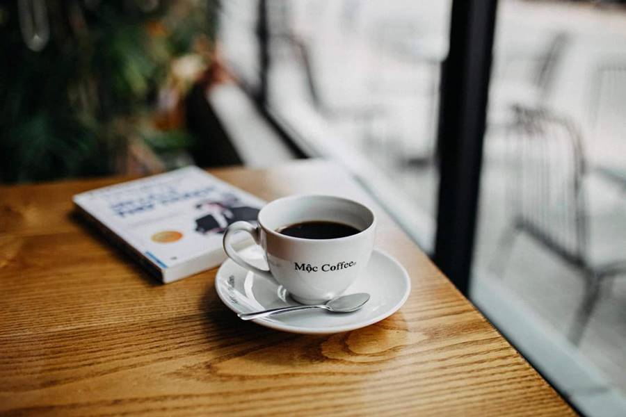 Tách cà phê trên bàn gỗ cạnh một quyển sách, nhìn ra cửa sổ với ánh sáng tự nhiên.