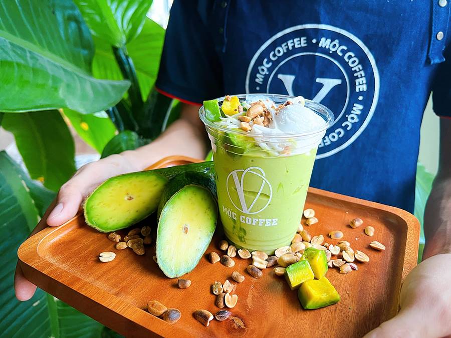 Ly sinh tố bơ trên khay gỗ, trang trí bằng hạt đậu phộng và miếng bơ tươi, được một người cầm.
