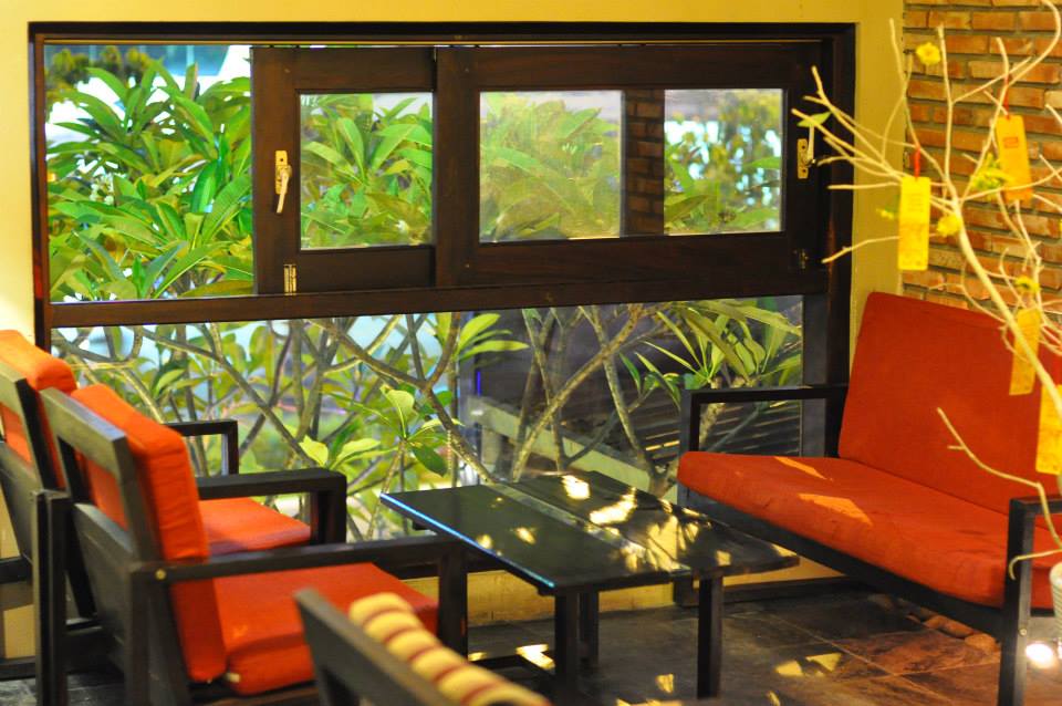 Khu vực ghế ngồi với b&agrave;n k&iacute;nh v&agrave; ghế đệm đỏ gần cửa sổ, nh&igrave;n ra c&acirc;y xanh b&ecirc;n ngo&agrave;i.