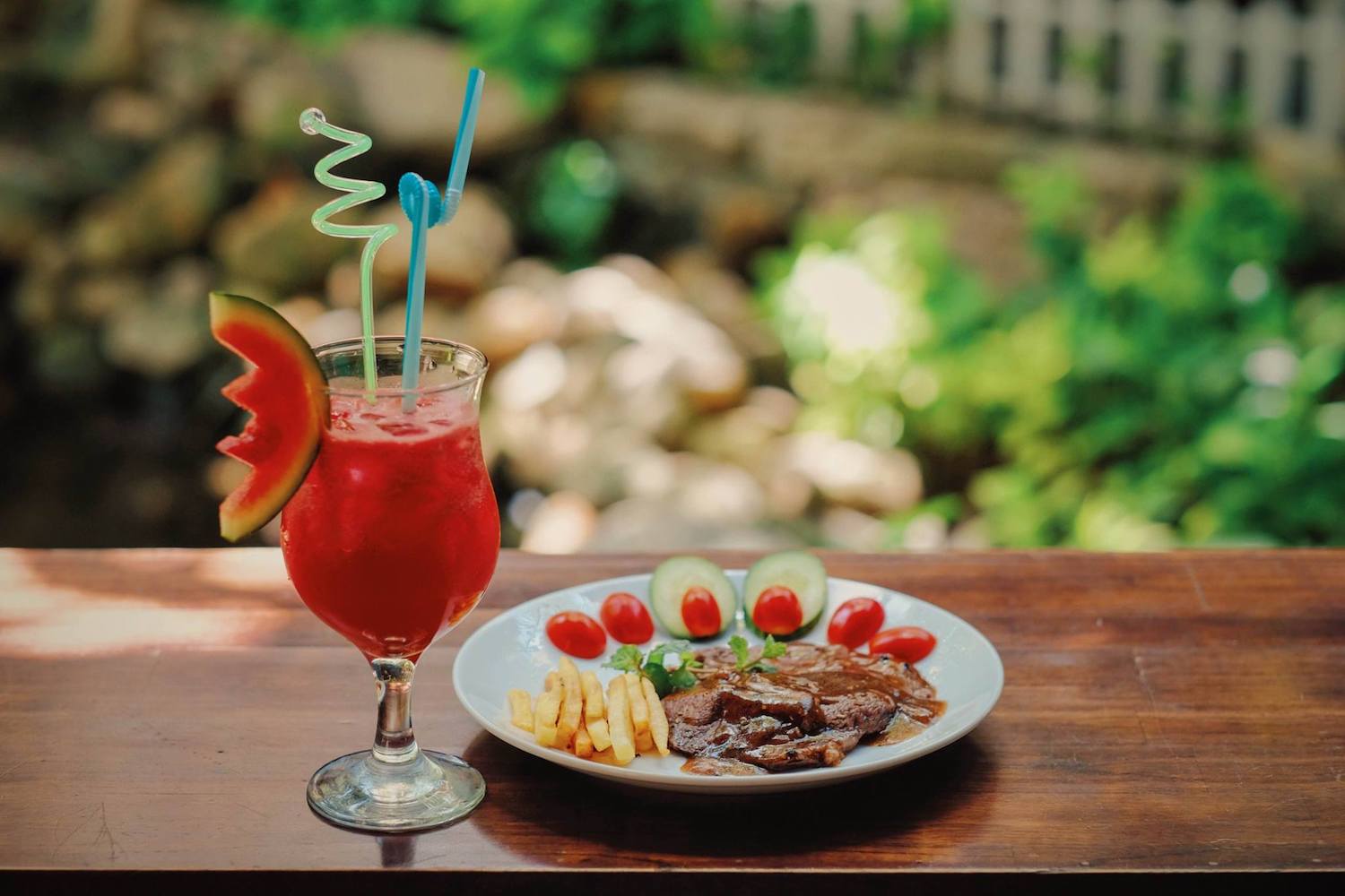 Ly nước đỏ với l&aacute;t dưa hấu v&agrave; ống h&uacute;t, b&ecirc;n cạnh đĩa thịt b&ograve;, khoai t&acirc;y chi&ecirc;n v&agrave; rau củ tr&ecirc;n b&agrave;n gỗ.