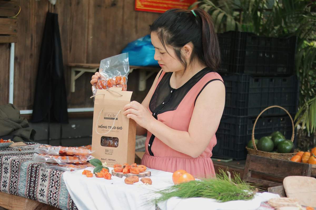 Người phụ nữ đang đ&oacute;ng g&oacute;i hồng sấy trong t&uacute;i giấy, tr&ecirc;n b&agrave;n c&oacute; nhiều g&oacute;i hồng sấy kh&aacute;c v&agrave; tr&aacute;i c&acirc;y.