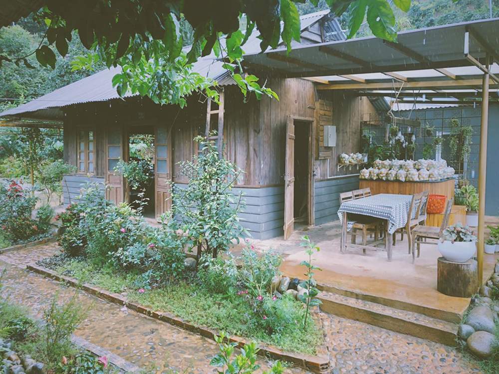 Ng&ocirc;i nh&agrave; gỗ nhỏ giữa khu vườn xanh, c&oacute; b&agrave;n ghế ngo&agrave;i trời v&agrave; nhiều c&acirc;y hoa xung quanh, trời đang mưa nhẹ.