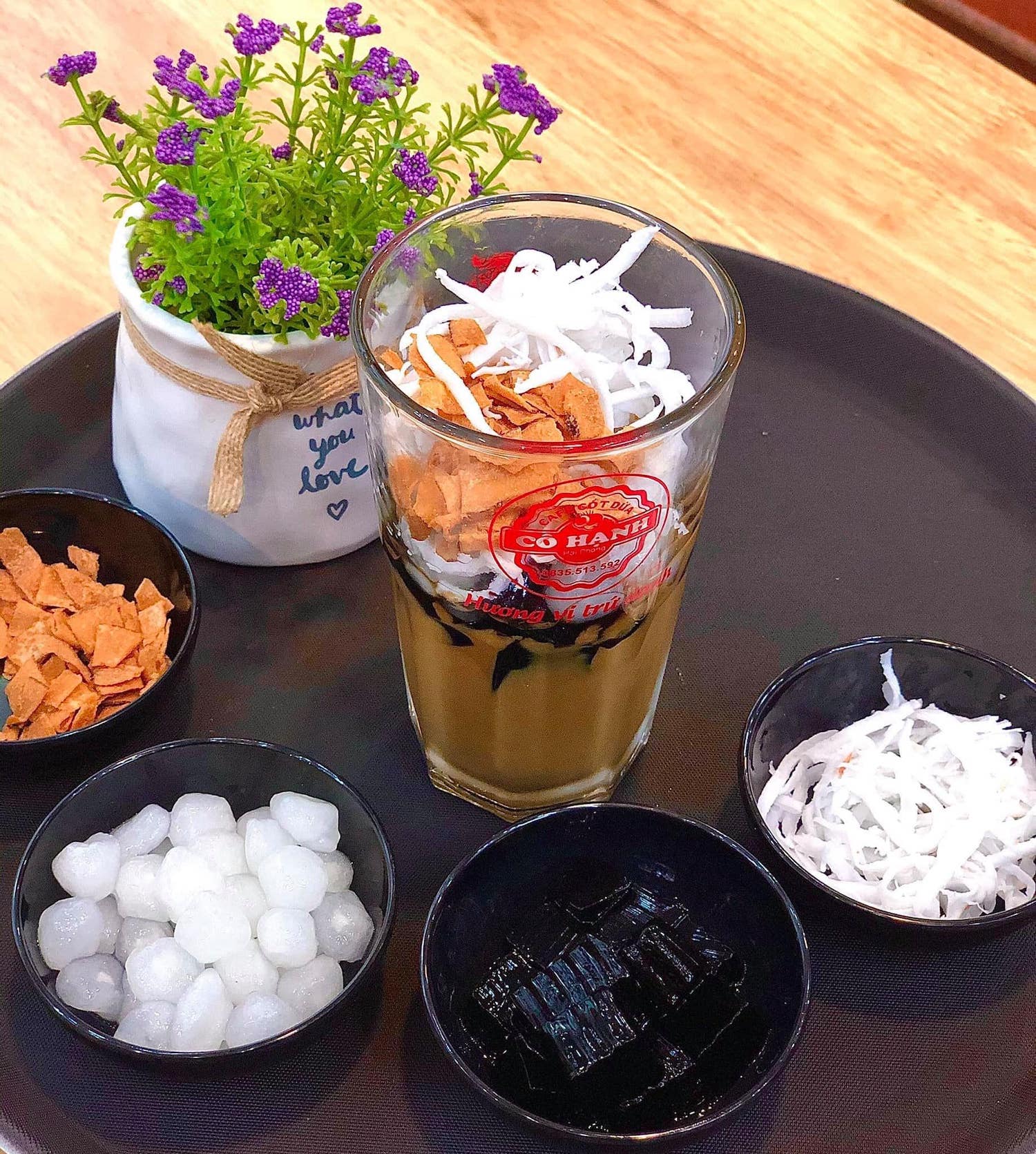 Ly c&agrave; ph&ecirc; sữa dừa với topping, trang tr&iacute; bởi chậu c&acirc;y nhỏ, đặt tr&ecirc;n khay tr&ograve;n m&agrave;u đen.