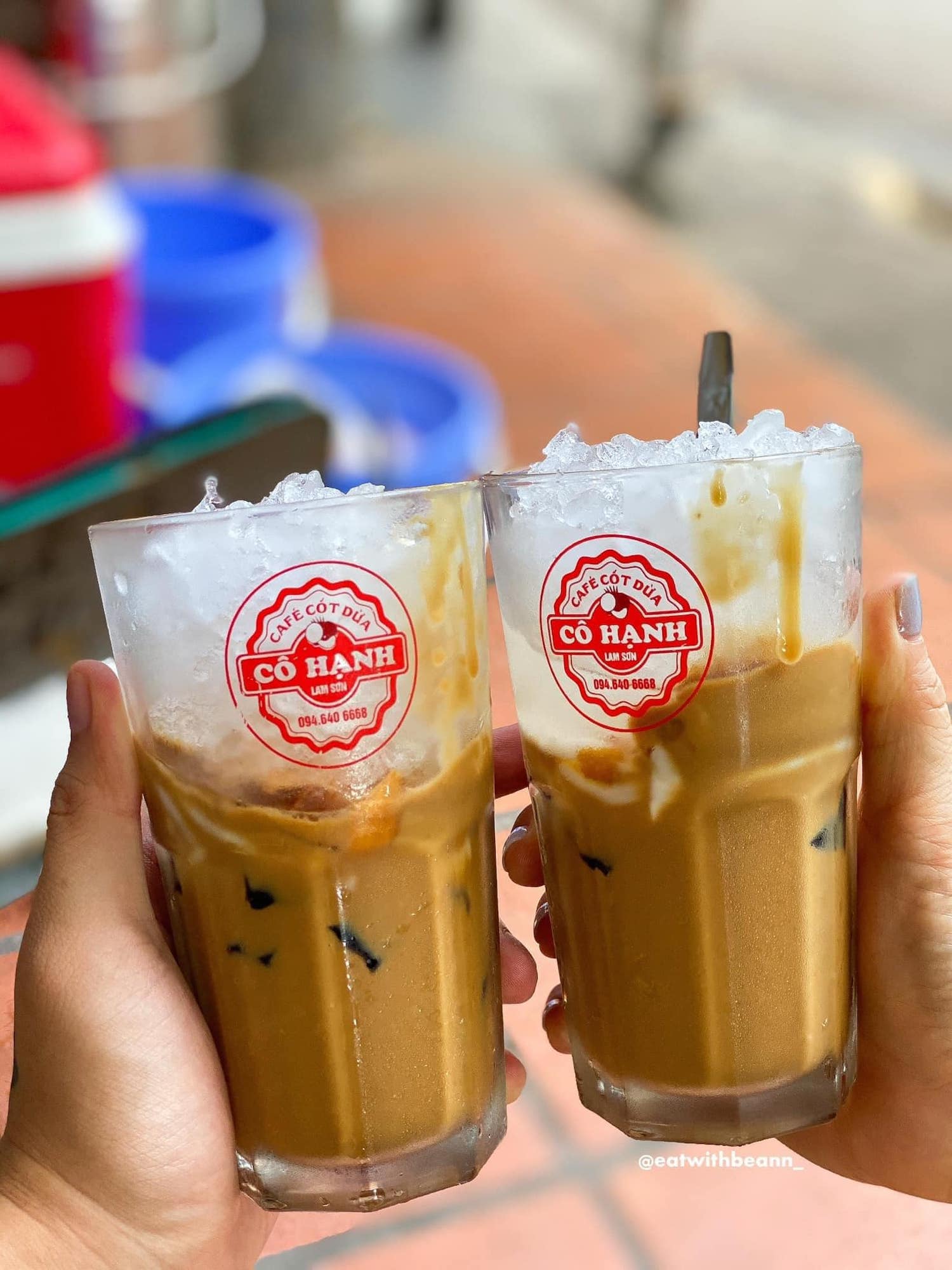 Hai ly c&agrave; ph&ecirc; cốt dừa đ&aacute; tr&ecirc;n tay người, nền mờ c&oacute; m&agrave;u sắc tươi s&aacute;ng v&agrave; v&agrave;i vật dụng kh&ocirc;ng r&otilde;.