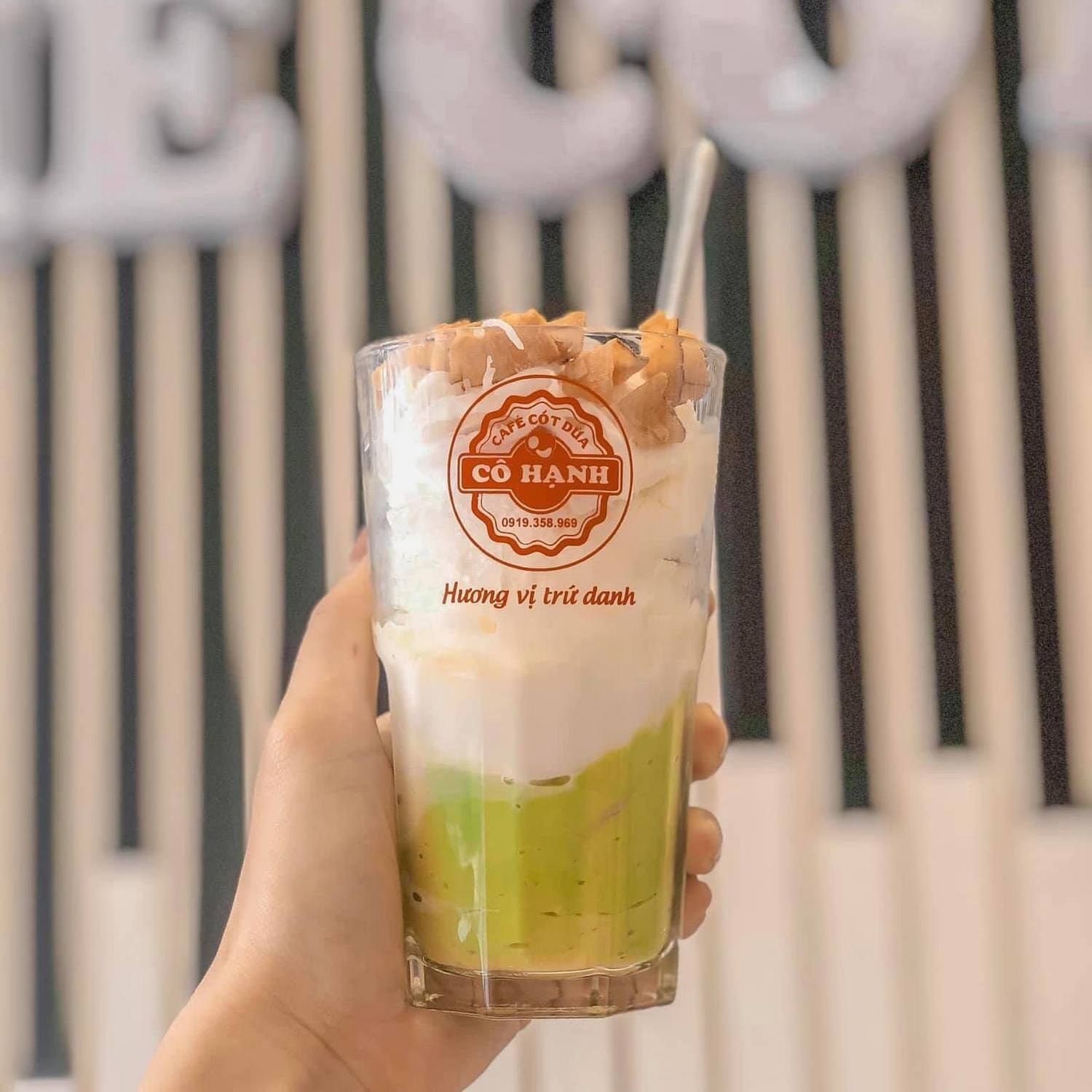 Ly sinh tố bơ dừa, với lớp kem, dừa b&agrave;o v&agrave; đ&aacute; b&agrave;o, được cầm tr&ecirc;n tay, nền sọc đen trắng.