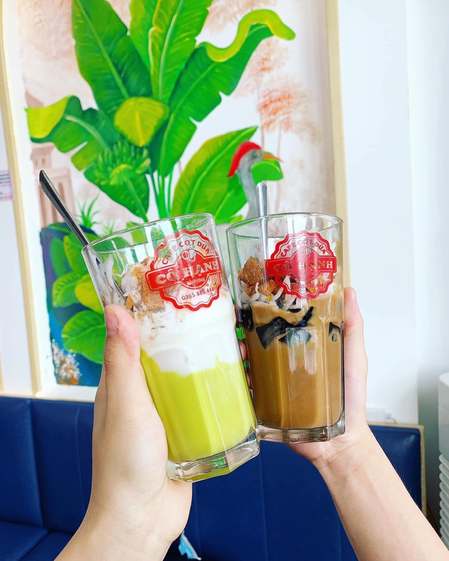 Hai ly c&agrave; ph&ecirc; cốt dừa với topping, được n&acirc;ng l&ecirc;n, nền l&agrave; bức tranh tường c&acirc;y xanh.