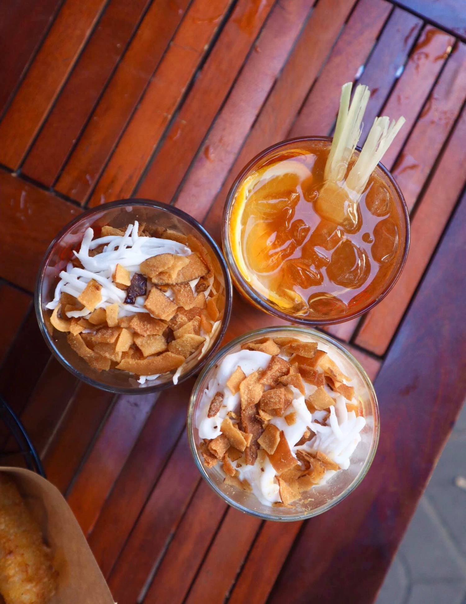 Ba ly đồ uống tr&ecirc;n b&agrave;n gỗ, hai ly c&oacute; lớp dừa v&agrave; b&aacute;nh, ly c&ograve;n lại c&oacute; l&aacute;t cam v&agrave; c&acirc;y xả.