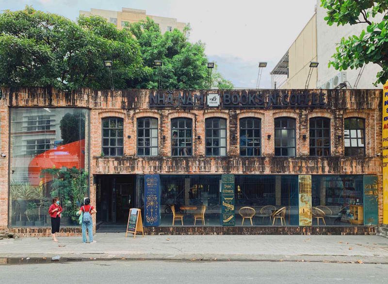 Hiệu sách và cà phê với mặt tiền gạch đỏ, cây xanh và hai người đứng ngoài trời.