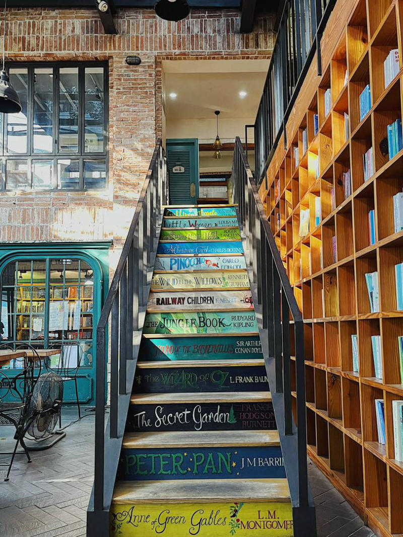 Cầu thang trang tr&iacute; bằng b&igrave;a s&aacute;ch nhiều m&agrave;u sắc, b&ecirc;n cạnh l&agrave; kệ s&aacute;ch gỗ trong kh&ocirc;ng gian qu&aacute;n c&agrave; ph&ecirc;.