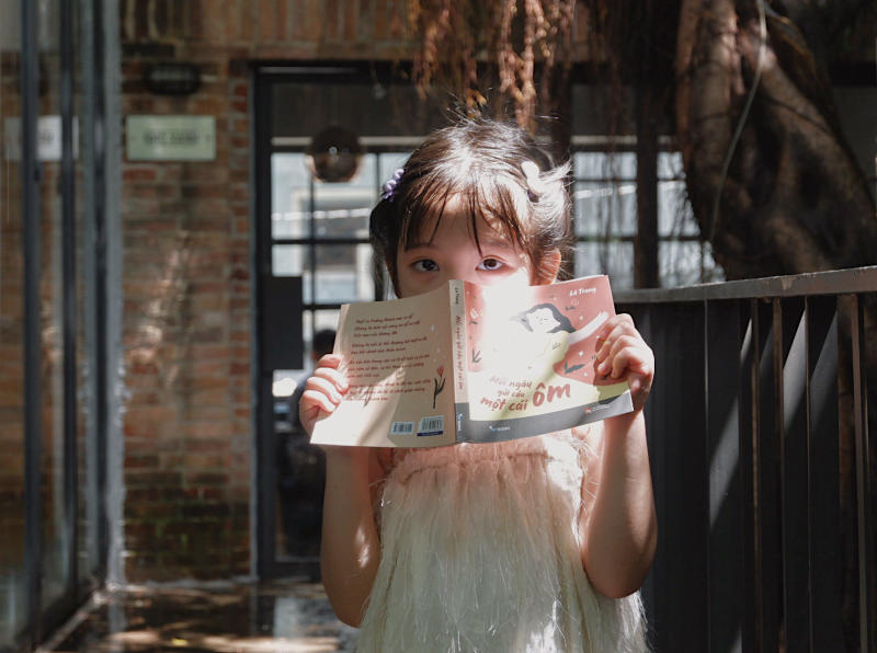 Em b&eacute; cầm cuốn s&aacute;ch, đứng trong kh&ocirc;ng gian c&oacute; &aacute;nh s&aacute;ng chiếu v&agrave;o, ph&ocirc;ng nền l&agrave; bức tường gạch.