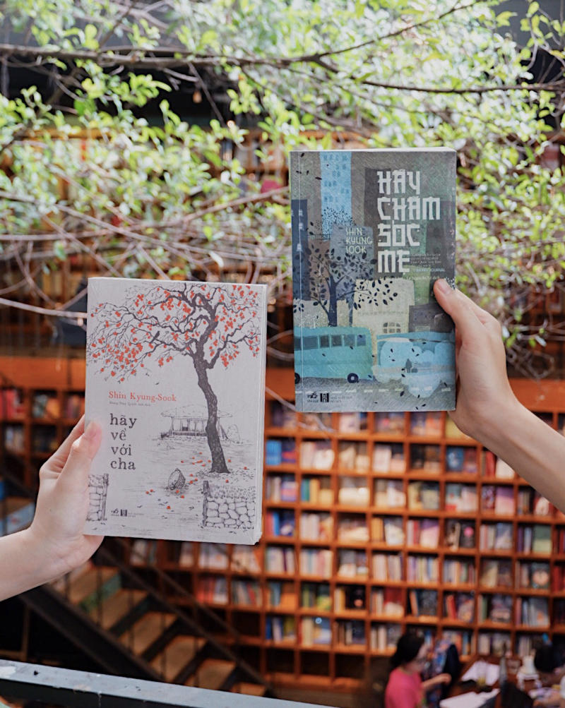 Hai người cầm hai cuốn s&aacute;ch trước gi&aacute; s&aacute;ch lớn trong kh&ocirc;ng gian thư viện với c&acirc;y xanh ph&iacute;a tr&ecirc;n.