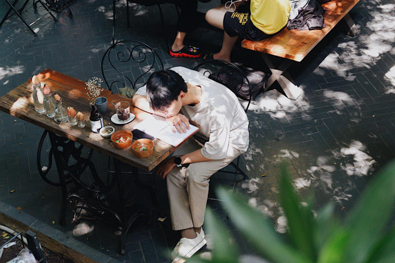 Người ngồi tại qu&aacute;n c&agrave; ph&ecirc; ngo&agrave;i trời, đọc s&aacute;ch tr&ecirc;n b&agrave;n gỗ, b&ecirc;n cạnh c&oacute; thức uống v&agrave; đồ ăn nhẹ.