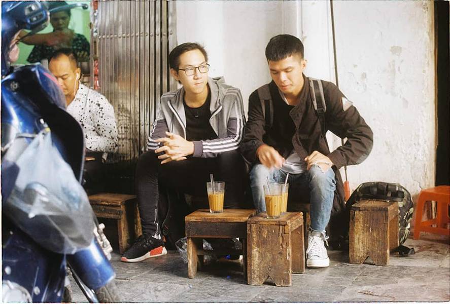 Hai người ngồi tr&ecirc;n ghế gỗ nhỏ, thưởng thức đồ uống tại một qu&aacute;n c&agrave; ph&ecirc; vỉa h&egrave;.