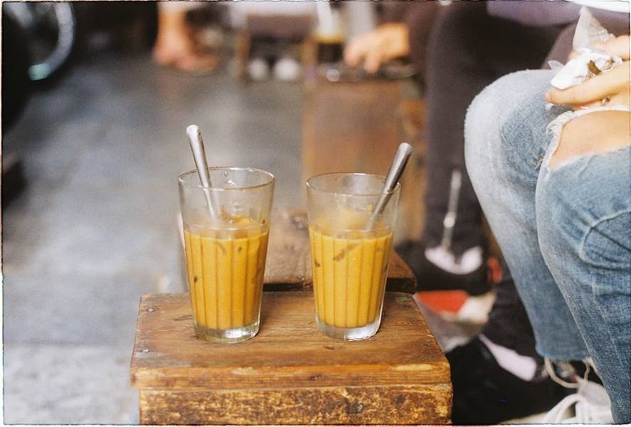 Hai ly c&agrave; ph&ecirc; sữa đ&aacute; tr&ecirc;n b&agrave;n gỗ, b&ecirc;n cạnh l&agrave; người mặc quần jeans r&aacute;ch ngồi.