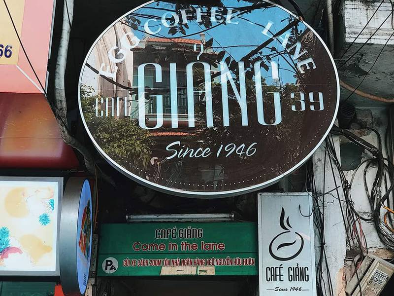 Biển hiệu qu&aacute;n c&agrave; ph&ecirc; với t&ecirc;n v&agrave; năm th&agrave;nh lập, xung quanh c&oacute; nhiều d&acirc;y điện v&agrave; biển quảng c&aacute;o kh&aacute;c.