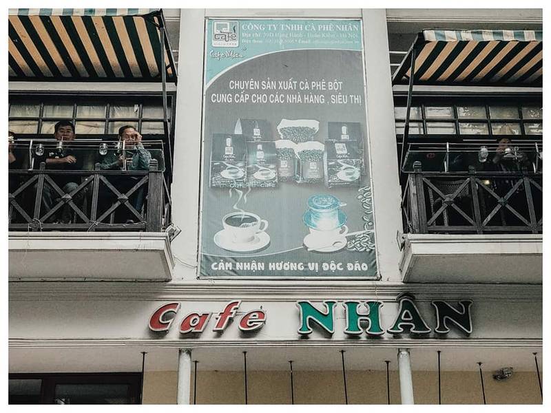 Mặt tiền qu&aacute;n c&agrave; ph&ecirc; với bảng hiệu lớn, c&oacute; người ngồi tr&ecirc;n ban c&ocirc;ng v&agrave; biển quảng c&aacute;o sản phẩm c&agrave; ph&ecirc;.